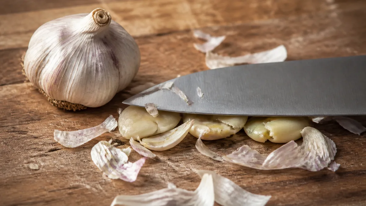 Zerdrückte Knoblauchzehen auf einem Holzbrett neben einer Knoblauchpresse, daneben eine bereitstehende Pfanne